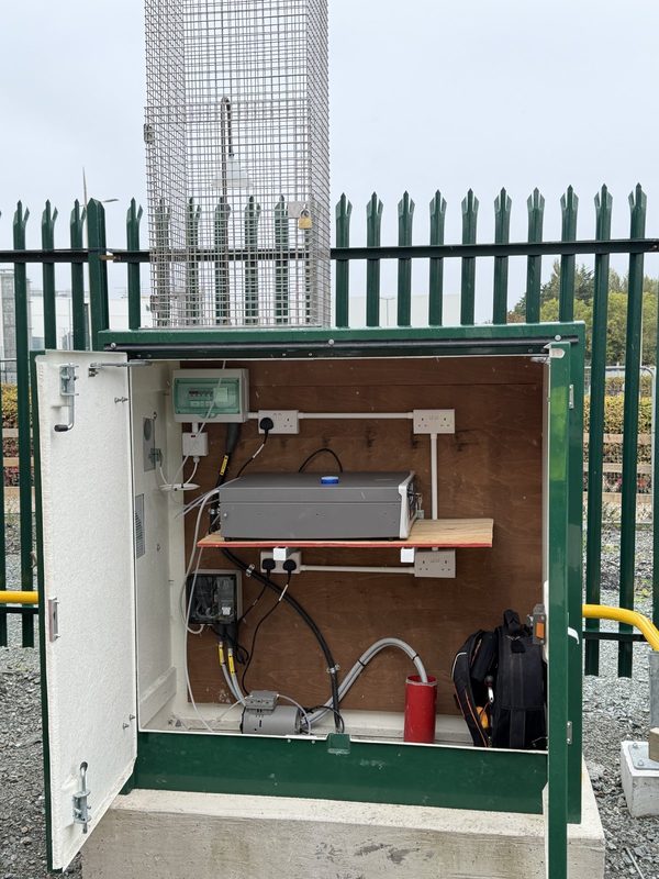 Open green air quality monitoring enclosure with an analyser on a shelf and a caged sample inlet above